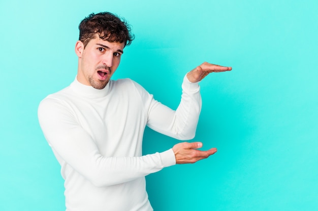 Young caucasian man isolated on blue wall shocked and amazed holding a copy space between hands.