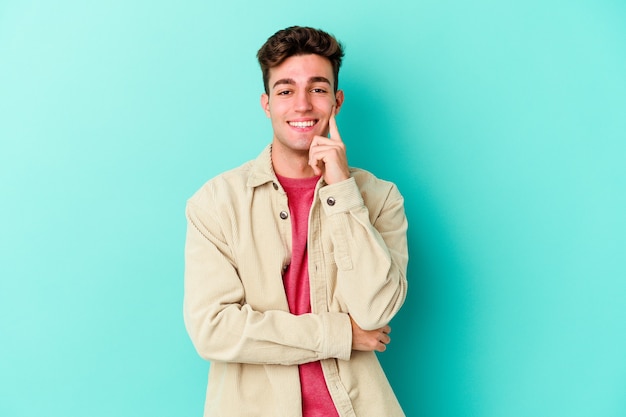 Young caucasian man isolated on blue wall smiling happy and confident, touching chin with hand.