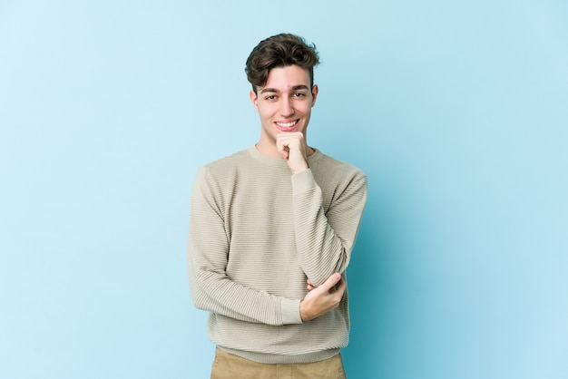 Young caucasian man isolated on blue wall smiling happy and confident, touching chin with hand.