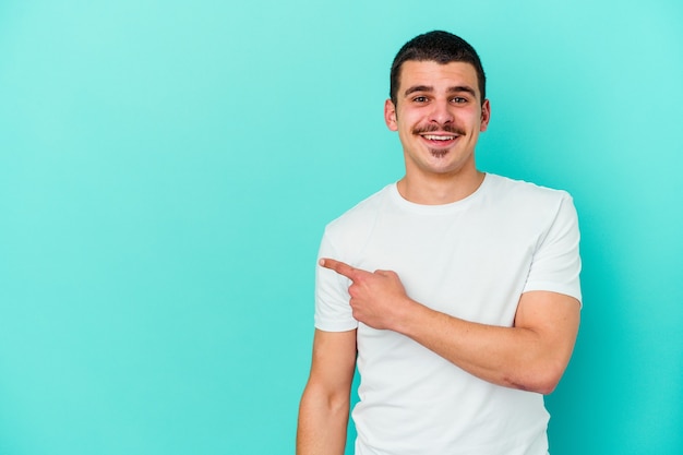 Young caucasian man isolated on blue wall smiling and pointing aside, showing something at blank space.