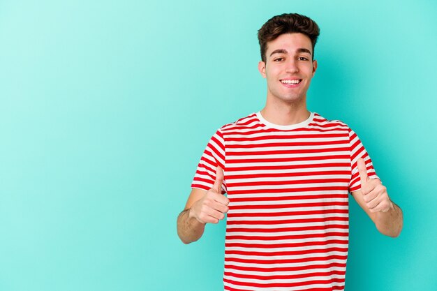 Young caucasian man isolated on blue wall smiling and raising thumb up