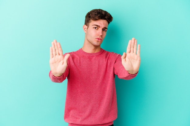 Young caucasian man isolated on blue wall standing with outstretched hand showing stop sign, preventing you.