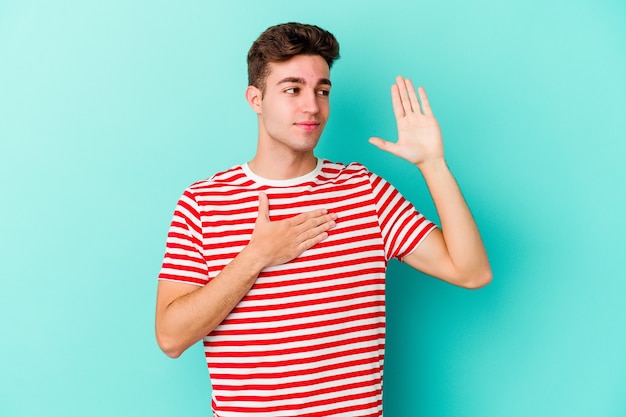 Young caucasian man isolated on blue wall taking an oath, putting hand on chest.