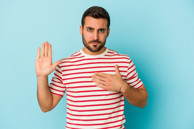 Young caucasian man isolated on blue wall taking an oath, putting hand on chest.