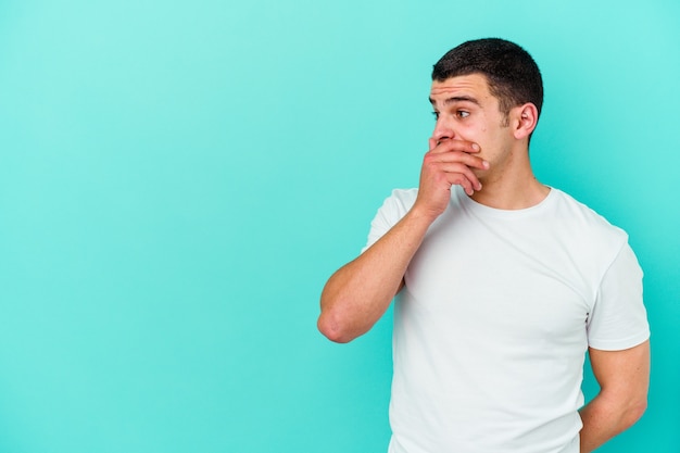 Young caucasian man isolated on blue wall thoughtful looking to a copy space covering mouth with hand