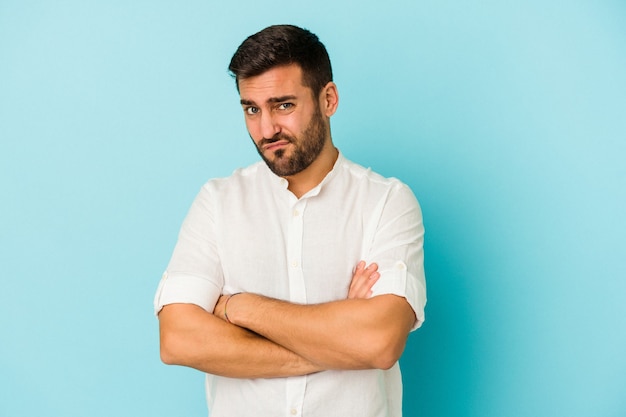 Young caucasian man isolated on blue wall unhappy looking at front with sarcastic expression