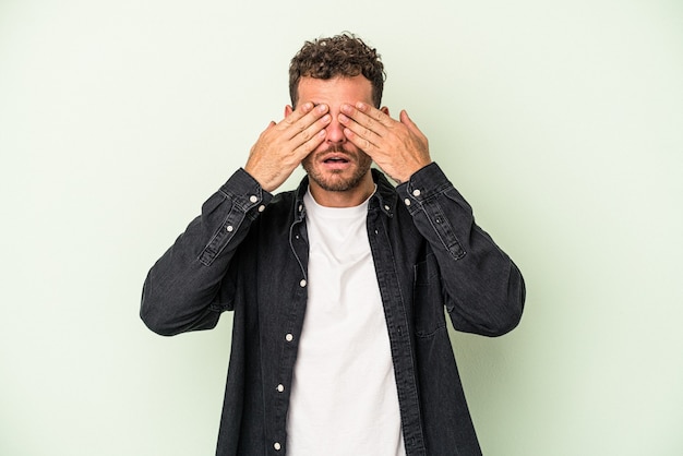 Young caucasian man isolated on green background afraid covering eyes with hands.