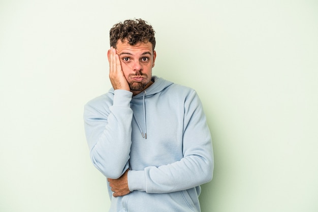 Young caucasian man isolated on green background blows cheeks, has tired expression. Facial expression concept.