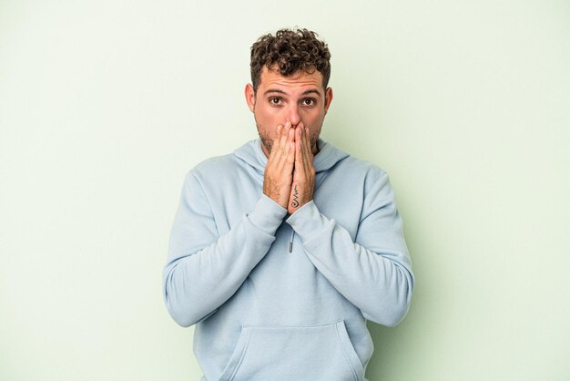 Young caucasian man isolated on green background covering mouth with hands looking worried.