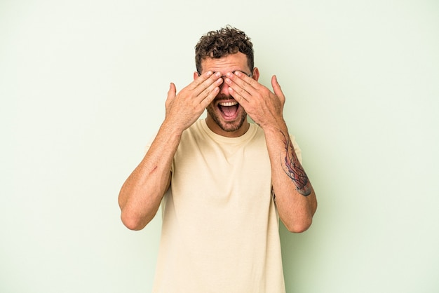 Young caucasian man isolated on green background covers eyes with hands, smiles broadly waiting for a surprise.