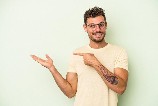 Young caucasian man isolated on green background excited holding a copy space on palm.