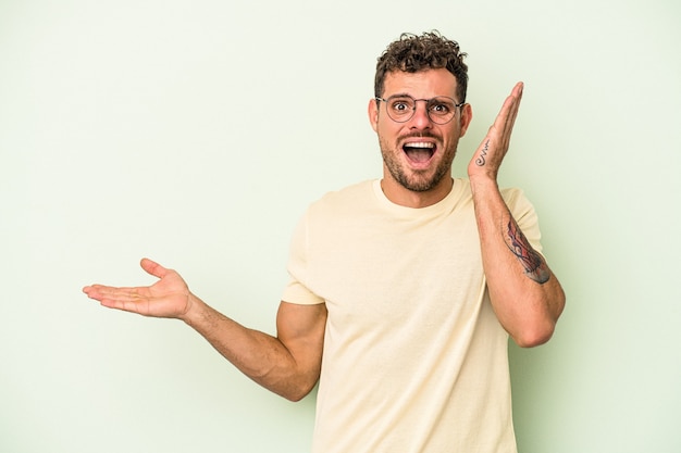 Young caucasian man isolated on green background holds copy space on a palm, keep hand over cheek. Amazed and delighted.