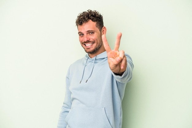 Young caucasian man isolated on green background joyful and carefree showing a peace symbol with fingers.