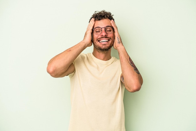 Young caucasian man isolated on green background laughs joyfully keeping hands on head. Happiness concept.