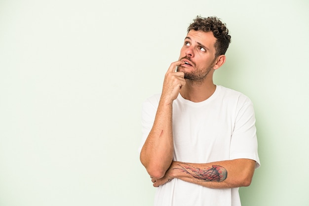 Young caucasian man isolated on green background looking sideways with doubtful and skeptical expression.