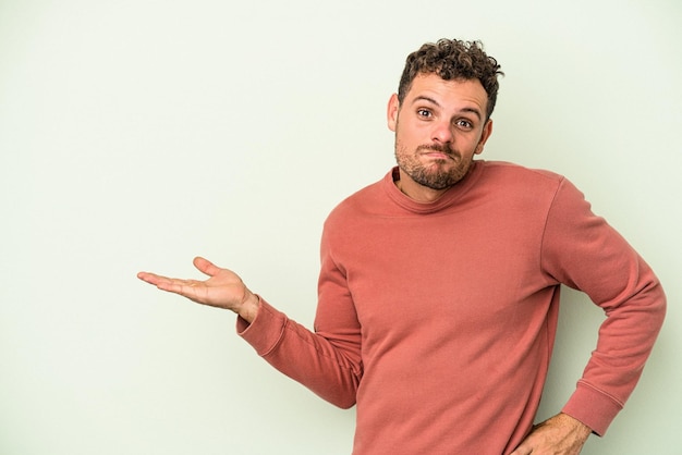 Photo young caucasian man isolated on green background showing a copy space on a palm and holding another hand on waist.