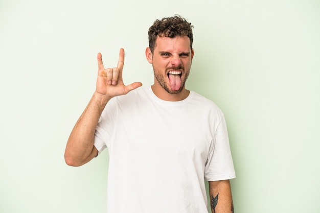 Young caucasian man isolated on green background showing a horns gesture as a revolution concept.