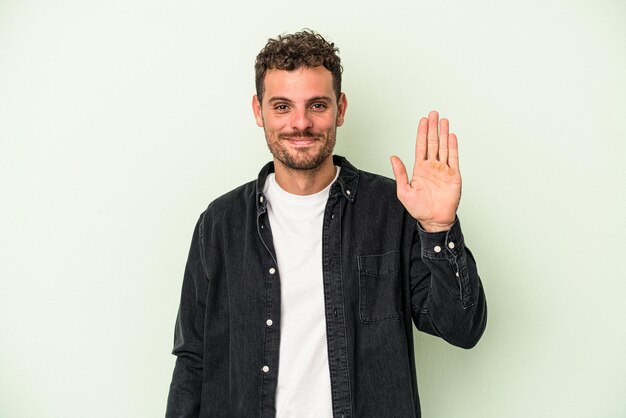 Young caucasian man isolated on green background smiling cheerful showing number five with fingers.