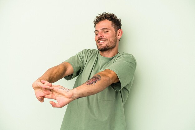 Young caucasian man isolated on green background stretching arms, relaxed position.