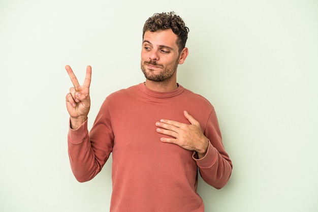 Foto giovane uomo caucasico isolato su sfondo verde prestando giuramento, mettendo la mano sul petto.