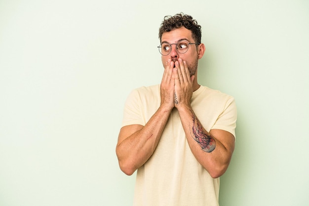 Young caucasian man isolated on green background thoughtful looking to a copy space covering mouth with hand.