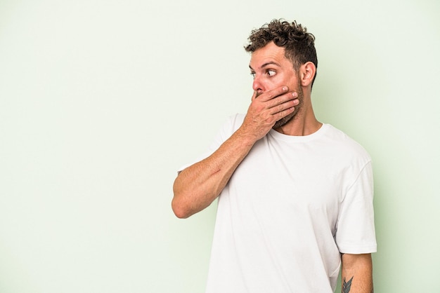 Young caucasian man isolated on green background thoughtful looking to a copy space covering mouth with hand.