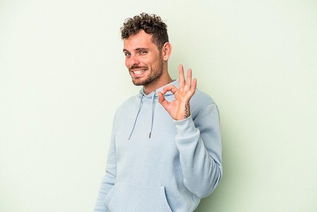 Young caucasian man isolated on green background winks an eye and holds an okay gesture with hand.