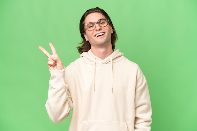 Young caucasian man isolated on green chroma background smiling and showing victory sign