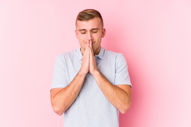 Young caucasian man isolated holding hands in pray near mouth, feels confident.