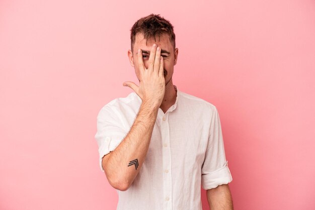 Young caucasian man isolated on pink background blink at the camera through fingers, embarrassed covering face.