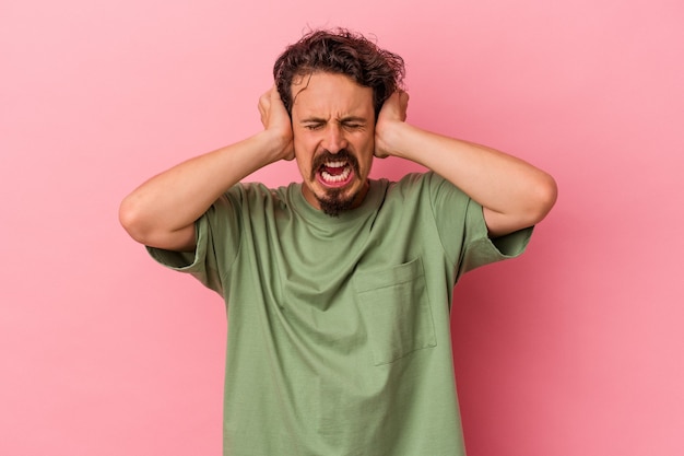 Young caucasian man isolated on pink background covering ears with hands trying not to hear too loud sound.