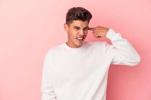 Young caucasian man isolated on pink background covering ears with hands.