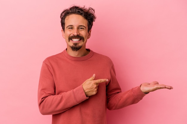 Young caucasian man isolated on pink background excited holding a copy space on palm.