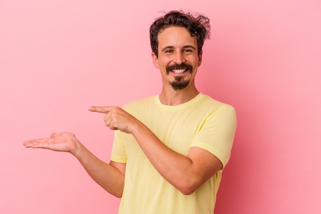 Young caucasian man isolated on pink background excited holding a copy space on palm.