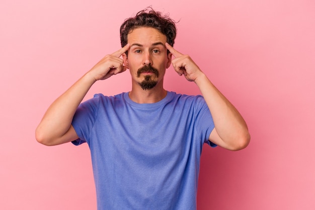Young caucasian man isolated on pink background focused on a task, keeping forefingers pointing head