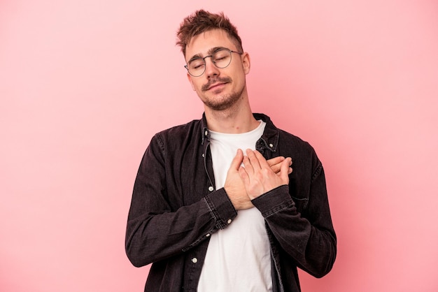 Young caucasian man isolated on pink background has friendly expression, pressing palm to chest. love concept.