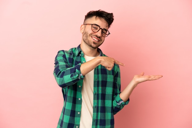 Young caucasian man isolated on pink background holding copyspace imaginary on the palm to insert an ad