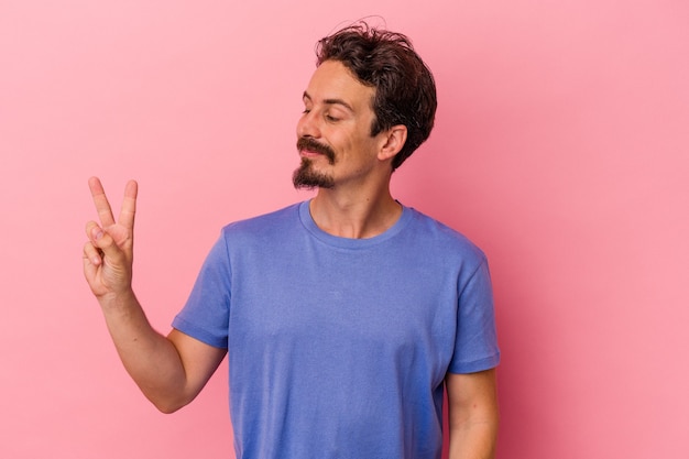 Young caucasian man isolated on pink background joyful and carefree showing a peace symbol with fingers.