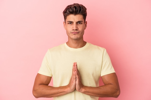 Young caucasian man isolated on pink background praying, showing devotion, religious person looking for divine inspiration.