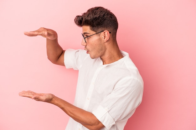 Young caucasian man isolated on pink background shocked and amazed holding a copy space between hands.