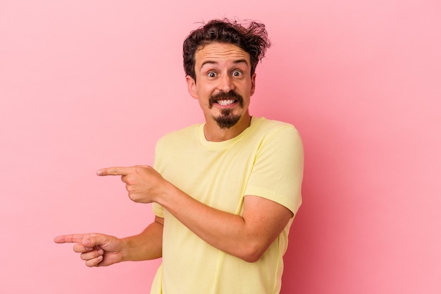 Young caucasian man isolated on pink background shocked pointing with index fingers to a copy space.