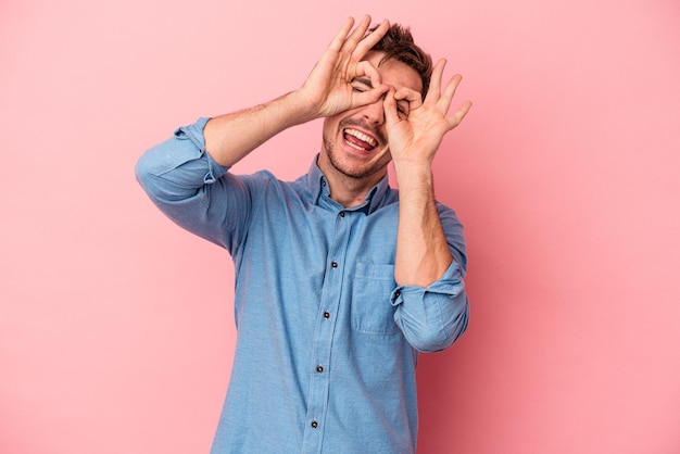 Young caucasian man isolated on pink background showing okay sign over eyes