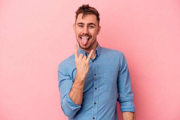 Young caucasian man isolated on pink background showing rock gesture with fingers