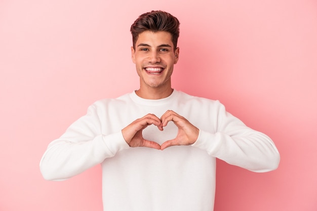 Young caucasian man isolated on pink background smiling and showing a heart shape with hands.