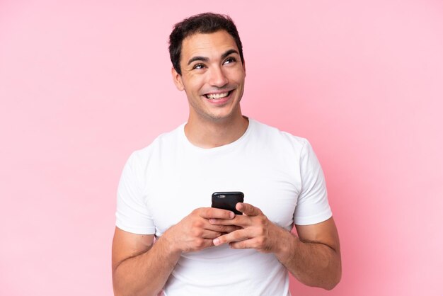 Young caucasian man isolated on pink background using mobile\
phone and looking up