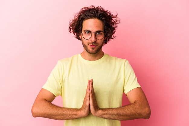 Young caucasian man isolated on pink bakcground praying, showing devotion, religious person looking for divine inspiration.