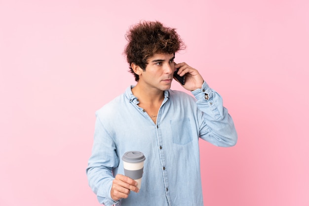 Photo young caucasian man over isolated pink wall holding coffee to take away and a mobile