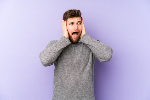 Young caucasian man isolated on purple background covering ears with hands trying not to hear too loud sound.