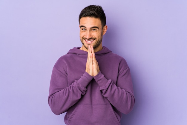 Young caucasian man isolated on purple background holding hands in pray near mouth, feels confidont.