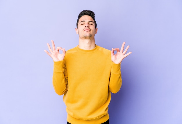 Young caucasian man isolated on purple background relaxes after hard working day, she is performing yoga.